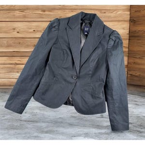 May include: A dark gray blazer with a single-button closure and a notched lapel. The blazer has a tailored fit with subtle shoulder padding and a small pocket on each side. The brand tag "GAP" is visible on the inside of the blazer.