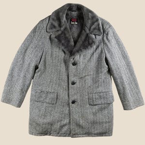 May include: A gray and white herringbone patterned coat featuring a dark gray faux fur collar and lining. The coat has three black buttons down the front, two side pockets with flaps, and a black label at the neck with the text Dan-Jac.