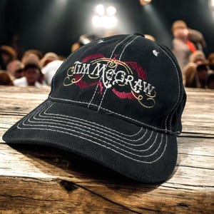 May include: A black baseball cap featuring white contrast stitching along the brim and panels. The front displays the text "Tim McGraw" embroidered in white, accented by a decorative red swirling design behind the lettering on the crown.