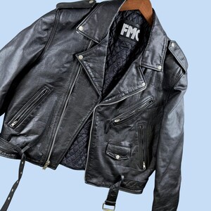 May include: A black leather motorcycle jacket featuring a quilted interior lining, silver-toned metal zippers, snap button lapels, shoulder epaulets, and a waist belt. The interior label displays the text FMC in white letters on a black background.