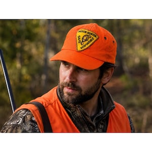 Puede incluir: Gorra de béisbol naranja brillante con un parche que dice "Oregon Hunter Safe Responsible". La gorra la lleva una persona que lleva un chaleco naranja y una chaqueta de camuflaje. El fondo es un bosque borroso.