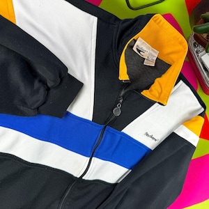 Könnte beinhalten: Eine schwarz-weiß-blau-gelbe Trainingsjacke mit Reißverschluss. Die Jacke hat ein Colorblock-Design mit einem weißen Streifen, einem blauen Streifen und einem gelben Kragen. Der Markenname "MacGregor" ist sichtbar.