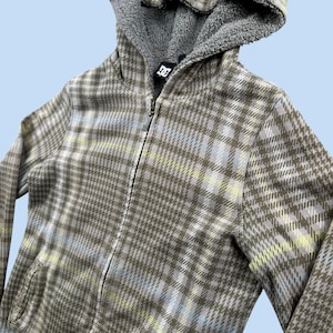 May include: A zip-up hooded sweatshirt featuring a brown, white, and light yellow plaid pattern. The garment includes a gray fleece-lined hood and a black DC brand logo tag at the neckline. The front has a full-length zipper closure.