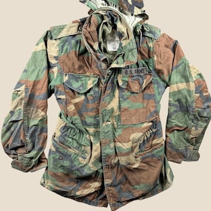 May include: A camouflage field jacket featuring a woodland pattern in shades of green, brown, and black. The jacket includes a high collar with a hood, a front zipper closure, four large button-flap pockets, and a black patch reading U.S. ARMY.