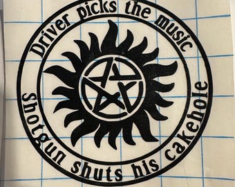 May include: Black sticker with the text "Driver picks the music, shotgun shuts his cakehole" in a circular design. The design includes a sunburst and a pentagram in the center. The sticker is on a grid-lined backing.