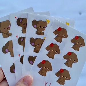 May include: Four sheets of stickers featuring brown cartoon bears with different expressions. Some bears have a red flower on their head, while others have a pink heart. The stickers are on a white background and the bottom sheet has the word "Yuls" written in pink.
