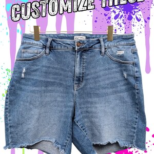 May include: Blue denim shorts with distressed details and a raw hem, hanging on a gold hanger. The text "CUSTOMIZE THESE" is displayed above the shorts against a purple and white background with paint splatters.