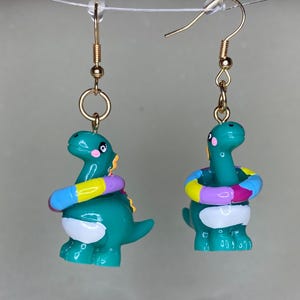 May include: A pair of whimsical earrings featuring teal dinosaur charms with pink cheeks and yellow accents. Each dinosaur wears a colorful inflatable ring. The earrings have gold-colored hooks.