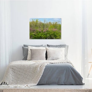 Op de afbeelding: Een rechthoekige wandkunstprint met een levendig boslandschap met felgroen gebladerte, paars bloeiende bomen en een helderblauwe lucht, opgehangen boven een bed met grijs beddengoed en een crèmekleurige gebreide sprei.