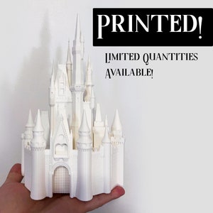 Puede incluir: Un modelo blanco impreso en 3D del Castillo de Cenicienta de Disney World. El castillo está detallado con torres, chapiteles y ventanas. El texto "PRINTED! LIMITED QUANTITIES AVAILABLE!" es visible en el fondo.
