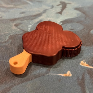 May include: A brown 3D printed clover-shaped object with a wooden handle. The object is on a blue and white patterned surface.
