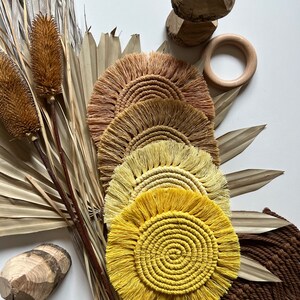 May include: Four round crocheted coasters with fringe. The coasters are in shades of yellow, orange, and brown. The coasters are arranged in a stack, with the largest coaster on the bottom and the smallest coaster on top.