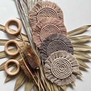 May include: Set of four round macrame coasters with fringe in shades of beige, brown, and pink. The coasters are arranged in a stack and are displayed on a background of dried palm leaves.