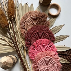 May include: A collection of handmade macrame coasters in shades of pink, brown, and tan. The coasters have a circular design with a fringed edge. The image also includes dried plants and wooden decorative elements, creating a natural, bohemian aesthetic.