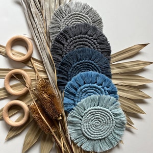 May include: Set of six round crocheted coasters in shades of blue and gray with fringe detailing. The coasters are arranged in a stack with the lightest color on top and the darkest color on the bottom. The coasters are displayed on a background of dried palm leaves.