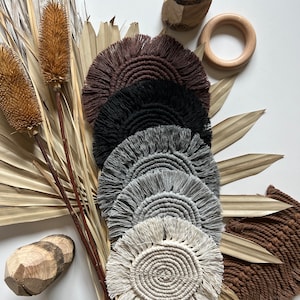 May include: A collection of handmade macrame coasters in various neutral colors, including brown, black, gray, and cream. The coasters have a circular design with a fringed edge. Decorative dried plants and wooden accents surround the coasters.