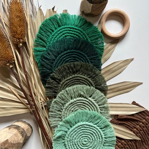 May include: A collection of handmade macrame coasters in shades of green, arranged in a fan shape. The coasters have a fringed edge and a spiral center. Dried palm leaves and wooden accents complement the coasters.