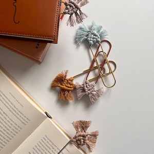 May include: Macrame bookmarks in various colors, including tan, blue, and mustard, are attached to paperclips and books. A brown leather journal with the name "Kaitlyn" is also visible. The bookmarks add a decorative touch to reading.