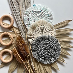 May include: Three handmade macrame coasters with fringe. The coasters are in a variety of colors and patterns, including white, beige, black, and blue. The coasters are arranged on a white background with dried palm leaves and brown stems.