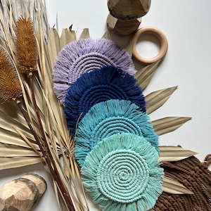 May include: A collection of handmade macrame coasters in shades of purple, blue, and teal. The coasters have a circular design with a fringed edge. The image also includes wooden rings, a carved wooden piece, and dried plants, creating a natural aesthetic.