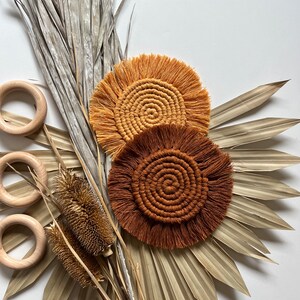 May include: Two round crocheted coasters with fringe, one in a burnt orange color and the other in a light orange color. The coasters are displayed on a background of dried palm leaves.