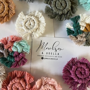 May include: A collection of handmade macrame coasters in various colors, including white, gray, teal, and pink. Each coaster features a spiral design with fringed edges. A business card with the text "Mackson & Odella" is also visible.