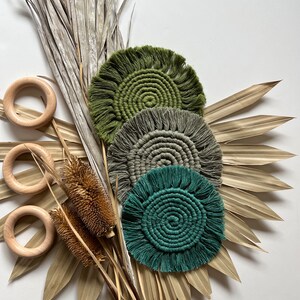 May include: Three macrame coasters in shades of green and gray, arranged with dried palm leaves and wooden rings. The coasters have a circular design with a fringed edge. The colors include olive green, gray, and teal.