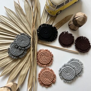 May include: A set of eight handmade macrame coasters in various colors, including gray, black, brown, and peach. The coasters are circular and feature a spiral design with fringe around the edges. The coasters are displayed on a white surface with a palm leaf and a toy van.