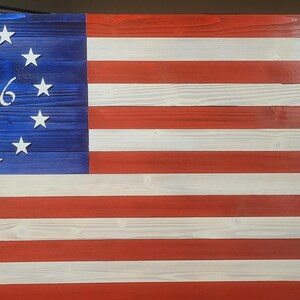 May include: A rectangular wooden wall art piece designed as a Betsy Ross flag with horizontal red and white stripes. The blue canton features thirteen white stars arranged in a circle surrounding the white painted text 1776 in the center.