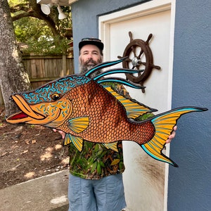 May include: A large, wooden fish sculpture painted in orange, yellow, and blue. The fish has a detailed design with scales and fins. The sculpture is being held by a person wearing a camouflage shirt and blue shorts.