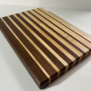 May include: A rectangular wooden cutting board with alternating dark brown and light brown stripes. The board has rounded edges and a smooth finish, suitable for kitchen use. The wood grain is visible, adding to its natural aesthetic.