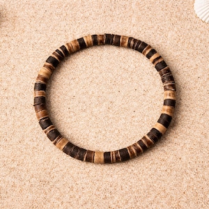 May include: A circular bracelet made of dark brown and tan wooden beads. The bracelet is resting on a bed of sand, with seashells scattered around it. The beads are cylindrical and arranged in alternating colors.