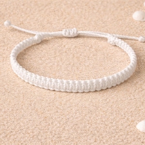 Peut inclure: Un bracelet macramé blanc avec une fermeture à nœud réglable. Le bracelet est fait de cordon tressé et est présenté sur une surface sablonneuse avec des coquillages. Un accessoire de plage simple.