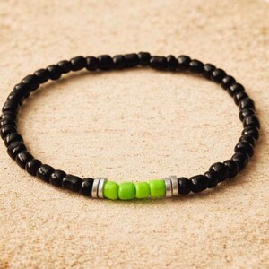 Op de afbeelding: Een zwart kralenarmband met een vleugje kleur. De armband heeft kleine, ronde zwarte kralen met een sectie van felgroene kralen en zilverkleurige afstandhouders. De armband wordt getoond op een zanderige ondergrond met schelpen.