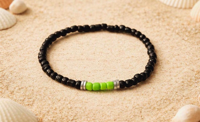 Op de afbeelding: Een zwart kralenarmband met een vleugje kleur. De armband heeft kleine, ronde zwarte kralen met een sectie van felgroene kralen en zilverkleurige afstandhouders. De armband wordt getoond op een zanderige ondergrond met schelpen.