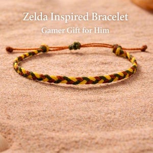 Puede incluir: Una pulsera trenzada con hilos amarillos, verdes y marrones, sobre un fondo arenoso. La pulsera tiene un cierre ajustable. El texto en la imagen dice "Zelda Inspired Bracelet" y "Gamer Gift for Him".