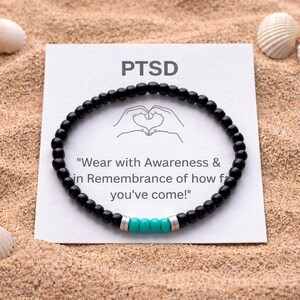 May include: Black beaded bracelet with turquoise accents, displayed on sand with seashells. A card with the word "PTSD" and the message "Wear with Awareness & in Remembrance of how far you've come!" is also present.