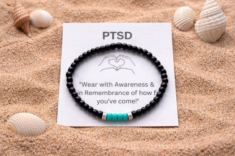 May include: Black beaded bracelet with turquoise accents, displayed on sand with seashells. A card with the word "PTSD" and the message "Wear with Awareness & in Remembrance of how far you've come!" is also present.