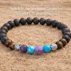May include: A Suicide Awareness Bracelet with black lava beads, brown tiger eye beads, and colorful accent beads. The bracelet includes the words "Support, Hope, Strength". The bracelet is displayed on a sandy surface.