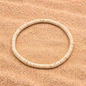 May include: A circular necklace made of small, light-colored beads. The necklace is resting on a textured, tan surface, creating a neutral and natural aesthetic. The beads appear to be uniform in size and shape.