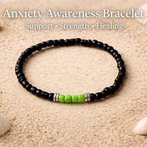 Op de afbeelding: Zwart kralenarmband met groene accentkralen en zilverkleurige afstandhouders. De armband ligt op zand met schelpen en de tekst "Anxiety Awareness Bracelet" erboven.