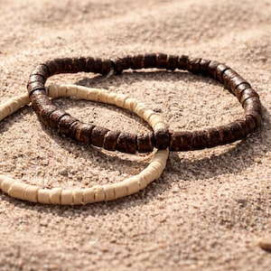 May include: Two beaded bracelets, one light beige and the other dark brown, rest on a sandy surface. The bracelets are made of small, cylindrical beads strung together. The dark brown bracelet is positioned over the beige one.