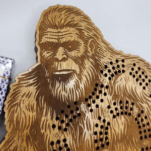 May include: Wooden game board shaped like a Bigfoot with a brown stain finish. The board has a black and white game path with numbers printed on it.
