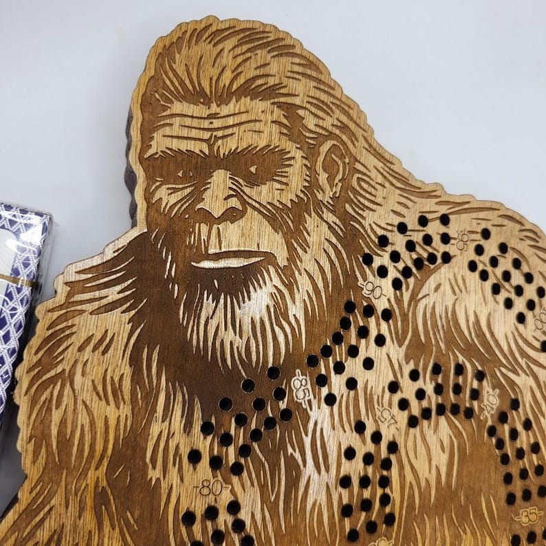 May include: Wooden game board shaped like a Bigfoot with a brown stain finish. The board has a black and white game path with numbers printed on it.