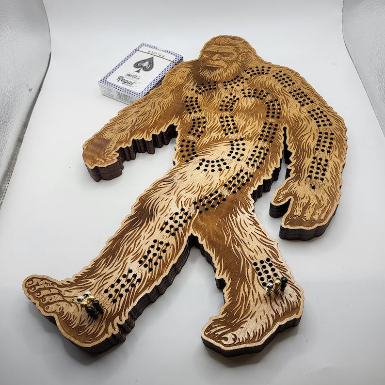 May include: A wooden game board shaped like a Bigfoot, with a brown stain finish. The board has a series of holes and paths for playing a game.