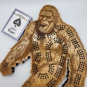 May include: A wooden game board shaped like Bigfoot, with a maze of holes and a playing card with the number 74 on it. The card is from a Regal deck of playing cards.