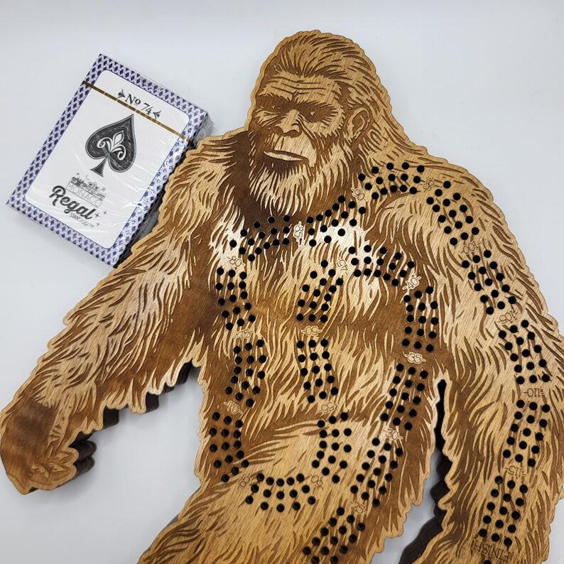May include: A wooden game board shaped like Bigfoot, with a maze of holes and a playing card with the number 74 on it. The card is from a Regal deck of playing cards.