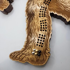 May include: A wooden Bigfoot game board with a brown stain. The board is shaped like a Bigfoot's foot. The game board has a track of holes with numbers from -15 to 15. The game board has a "START" label and a few game pieces on the board.