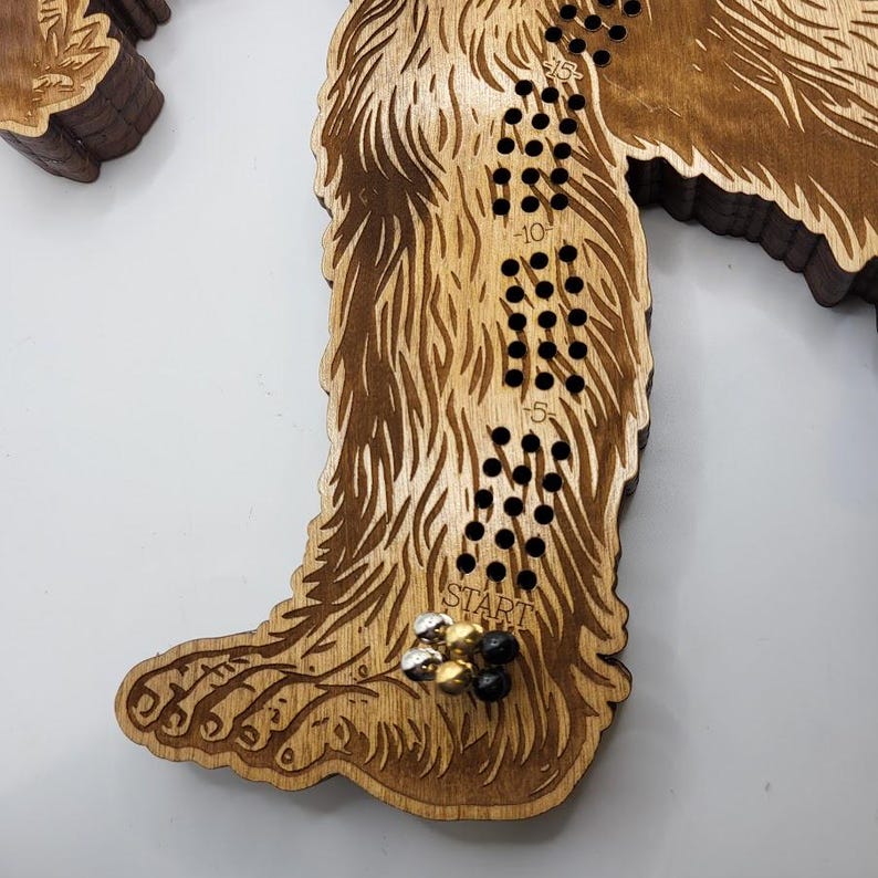 May include: A wooden Bigfoot game board with a brown stain. The board is shaped like a Bigfoot's foot. The game board has a track of holes with numbers from -15 to 15. The game board has a "START" label and a few game pieces on the board.