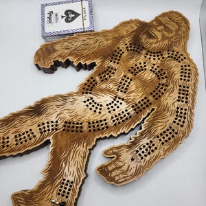 May include: A wooden Bigfoot game board with a brown stain. The board is shaped like a Bigfoot with a detailed design of fur and footprints. The game board has a series of holes for playing the game.